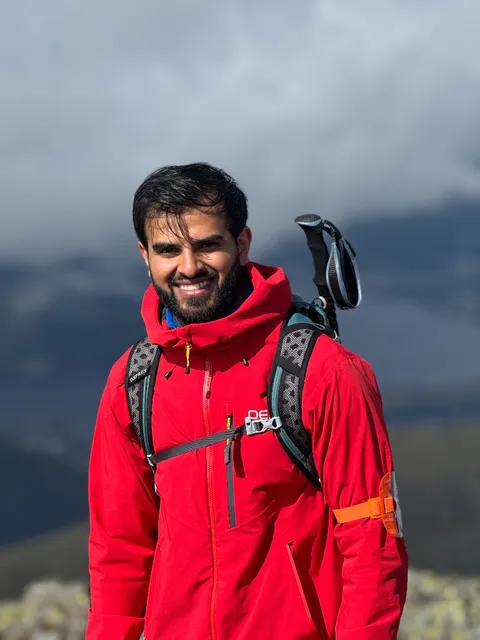 Afzal M. smiling in a red hiking jacket, with a cloudy mountain range behind him.